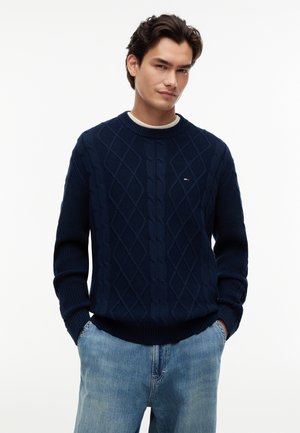 Junger Mann mit dunklen Haaren, der einen marineblauen Zopfpullover und hellblaue Jeans trägt, die Hände in den Taschen, schaut leicht seitlich.
