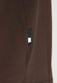 Close-up van bruin gebreid stof met een klein zwart-wit rechthoekig label dat langs een verticale naad is genaaid.