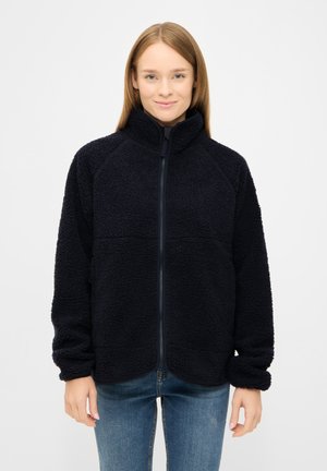 Giovane donna con capelli lisci di colore castano chiaro, indossa una giacca in pile nera con zip e jeans blu, in piedi su uno sfondo bianco semplice.