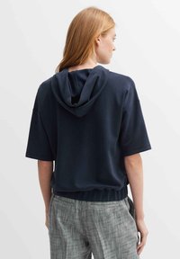 Felpa navy con cappuccio, maniche corte e orlo elastico. Abbinata a pantaloni grigi texturizzati, mostrata di lato.