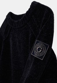 Schwarzer Pullover mit strukturiertem Gewebe und geripptem Rundhalsausschnitt, ausgestattet mit einem rechteckigen Logopatch mit blauen und beigen Akzenten am Ärmel.