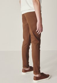 Pantalon en coton marron à coupe slim, ourlets retroussés, accompagné de baskets montantes en lacets brun foncé. Le tissu semble lisse avec une texture minimale.