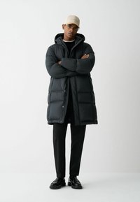 Črna puhasta bunda s kapuco, ki ima prešite dele in zapiranje na gumbke. Kombinirana s črnimi hlačami in črnimi čevlji, stoji ob enotni ozadju.