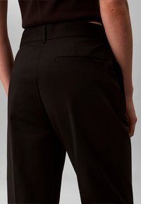 Nahaufnahme einer Person, die schwarze, maßgeschneiderte Hose mit einer Leistentasche am Rücken und schwarzem Oberteil trägt, von hinten auf Taillen- und Hüfthöhe betrachtet.