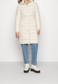 Kurtka puchowa w kremowym kolorze z odpinanym futrzanym kapturem, charakteryzująca się pikowanym wzorem i zapięciem na zamek błyskawiczny. W połączeniu z jasnoniebieskimi dżinsami i białymi botkami.