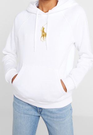 Osoba w białej bluzie z kapturem z złotym logo gracza polo i niebieskich dżinsach, z rękami wsuniętymi w przednią kieszeń, na jednolitym tle.