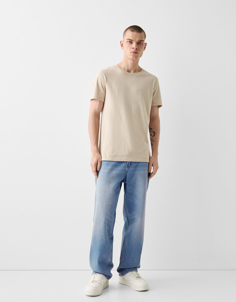 Beige Baumwoll-T-Shirt mit Rundhalsausschnitt, kombiniert mit hellblauen Jeans mit geradem Bein und weißen Sneakers. Minimalistisches Design, ohne Muster.