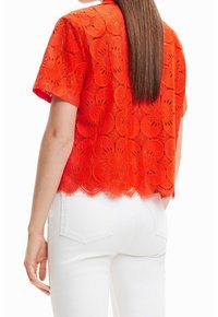 Top rouge à manches courtes brodé avec un motif en dentelle florale, ourlet festonné et coupe décontractée, associé à un jean blanc.