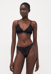 Zwarte kanten lingerie set met een bralette voorzien van geschulpte randen en een elastische band met merknaam, samen met een bijpassende kanten string.