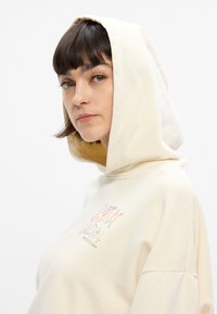 Κρεμ hoodie με μαλακή υφή, διαθέτοντας γραφιστικό σχέδιο και αστέρια μπροστά, συνδυασμένο με ρυθμιζόμενη κουκούλα.