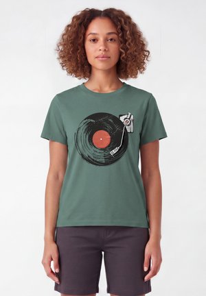 Femme aux cheveux bouclés portant un t-shirt vert avec un graphisme de disque vinyle noir et rouge et platine, associé à un short foncé.
