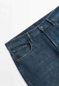 Modré džíny z denimu s přední kapsou na minci, kovovými nýty, poutky na pásek a knoflíkovým zapínáním na bílém pozadí.