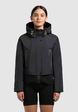 Naisel mustas lühikeses kapuutsiga jopes kollaste nööridega ja mustade rattapükstega, seisab ühevärvilise tausta ees.