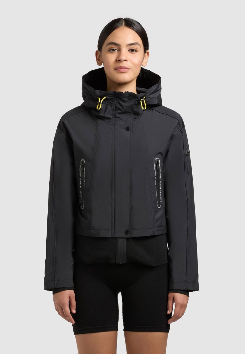 Femme portant une veste à capuche noire courte avec des cordons jaunes et un short de cyclisme noir, debout devant un fond uni.