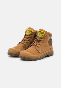 Botas de tobillo de ante marrón con una puntera de goma, suela texturizada, acentos de logo amarillos y verdes, y cremallera lateral para poner y quitar fácilmente.