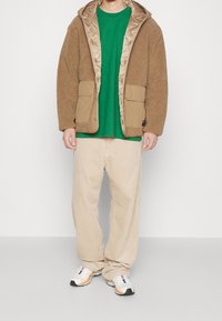 Chaqueta de forro polar marrón con dos bolsillos frontales, pantalones de pana beige y zapatillas blancas con detalles naranjas, combinados con una camisa verde debajo.