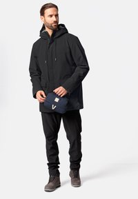 Zwarte waterdichte jacket met capuchon, uitgerust met twee voorzakken, gedragen met zwarte broek en grijze wandelschoenen; vasthouden aan een marineblauwe tas met een logo.