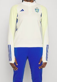 Maglia atletica a maniche lunghe color crema con spalle giallo chiaro, dettagli blu e logo Adidas. Presenta un collo con zip a un quarto e una vestibilità elegante.