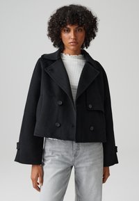 Cappotto corto nero in materiale tessuto, con rever oversize, bottoni doppi e maniche ampie, indossato sopra un top bianco.