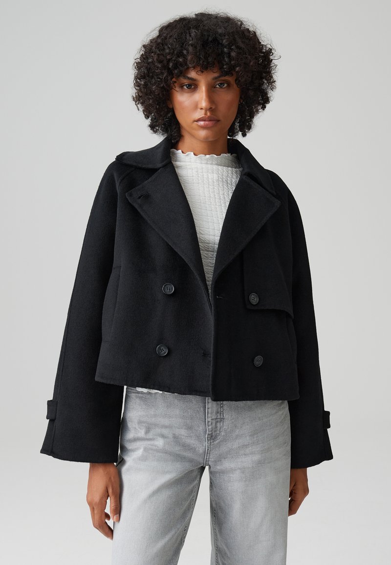 Cappotto corto nero in materiale tessuto, con rever oversize, bottoni doppi e maniche ampie, indossato sopra un top bianco.