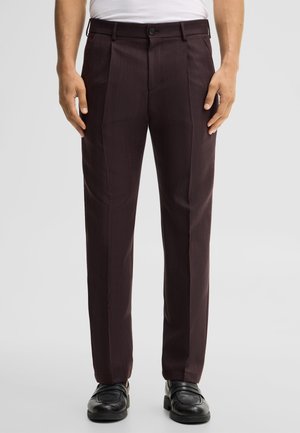 Mężczyzna ubrany w ciemnobrązowe, dopasowane spodnie i czarne skórzane eleganckie buty, stojący na jednolitym jasnym tle.