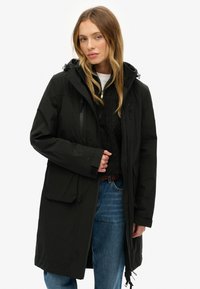 Femme portant un long manteau noir à capuche sur un pull en maille noire et un jean bleu, se tenant contre un fond blanc uni.