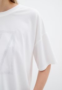 Valge lühikeste varrukatega T-särk, millel on suur metallist nupukestest koosnev ‚N‘ muster, lõdva istuvuse ja pehme tekstuuriga.