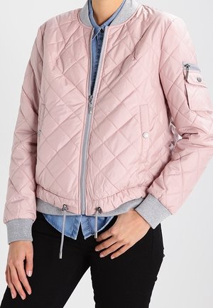 Person trägt eine hellrosa gesteppte Bomberjacke mit grauen Bündchen und Saum über einem blauen Hemd und schwarzen Hosen.