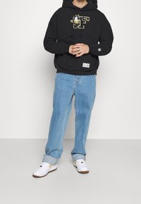 Černá mikina s kapucí s grafickým designem, modré džínové kalhoty a bílé tenisky s hnědými detaily. Volný střih a ležérní styl.