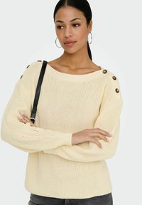 Femme portant un pull en maille crème avec des boutons aux épaules, de grandes boucles d'oreilles anneaux, et une sangle noire sur l'épaule gauche, bras croisés.