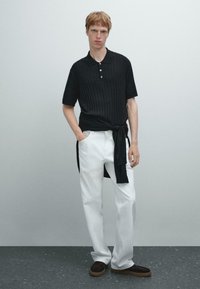 Polo shirt nero a coste con tre bottoni, pantaloni bianchi a gamba larga e scarpe marroni scure. Un maglione nero legato in vita.