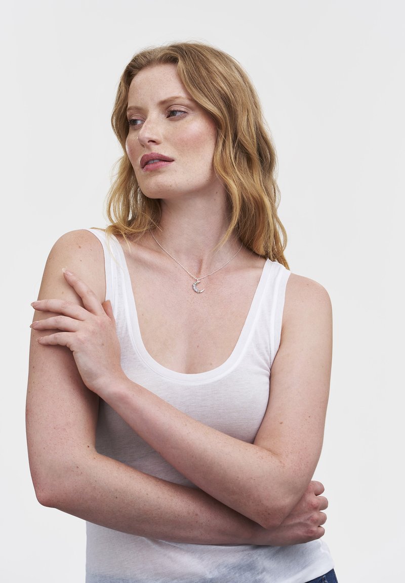 Pikajuustega punapea naine, kes kannab valget topi ja poolkuu kaelakeed, ristab käed ning vaatab küljele ühevärvilise tausta ees.