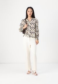 Krämfärgad blus med mönster i marinblåa diamantformer, tillsammans med vita texturerade vidbyxor och vita sandaler med blockklack. Brun handväska.