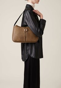Brown textured handbag with a rounded shape, featuring a gold circular clasp and a long shoulder strap, carried by a person in a black leather jacket.