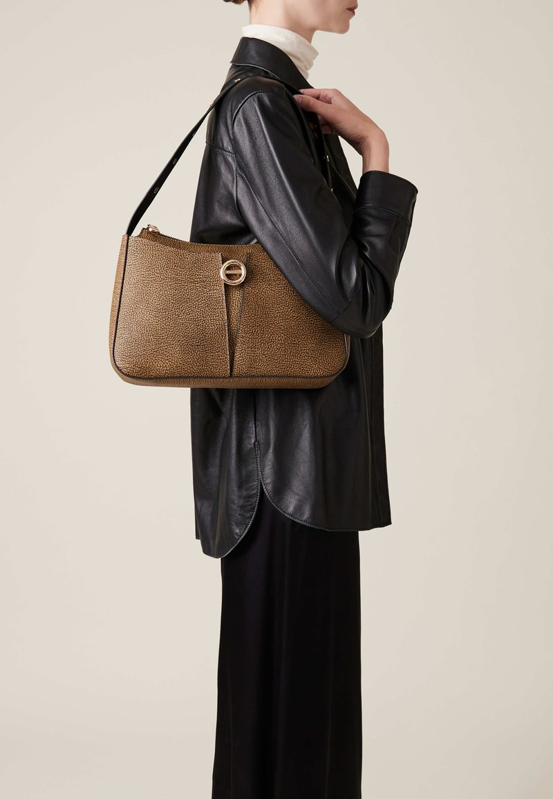 Brown textured handbag with a rounded shape, featuring a gold circular clasp and a long shoulder strap, carried by a person in a black leather jacket.