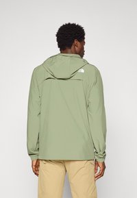 Mand iført en lysegrøn hættetrøje og beige bukser, vist bagfra mod en ensfarvet baggrund.