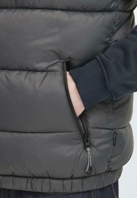 Grau gepolsterte Weste mit Reißverschlusstaschen, gerippten Bündchen und glatter Textur. Auffälliger Farbkontrast mit dem navyblauen Stoff an den Bündchen.