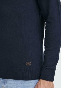 Dunkelblauer Strickpullover mit gerippten Manschetten und Saum, ausgestattet mit einem Lederetikett mit geprägtem Text an der Unterseite. Weiche Textur und figurbetontes Design.
