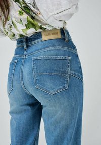 Vaqueros de mezclilla azul claro con pernera recta, que cuentan con bolsillos traseros y una etiqueta de cuero marrón claro marcada con "Salsa Jeans" en la cintura.