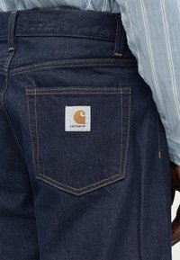 Οπίσθια όψη ατόμου που φορά σκούρα μπλε τζιν Carhartt με ορατό λογότυπο της μάρκας στην πίσω τσέπη και μανίκι πουκάμισου με ανοιχτό μπλε ριγέ μοτίβο.