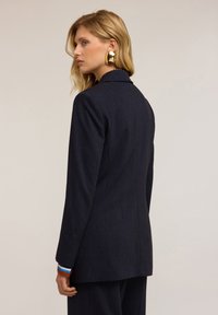 Granatowa marynarka wykonana z teksturowanej tkaniny, o dopasowanym kroju, z wyrazistymi klapami i kontrastowymi mankietami. Minimalistyczne detale w całej konstrukcji.