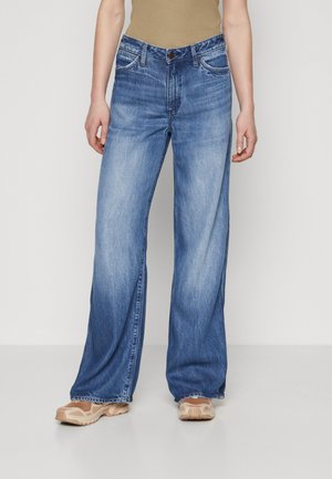 Άτομο που φορά ανοιχτό μπλε τζιν παντελόνι με φαρδιά μπάτζα, μπεζ τοπ και μπεζ αθλητικά παπούτσια, στέκεται μπροστά σε ένα απλό λευκό φόντο.