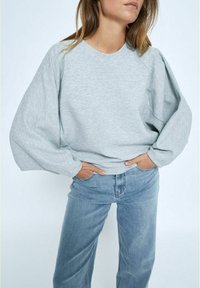 Helle graue, strukturierte Sweatshirt mit voluminösen Ärmeln, Rundhalsausschnitt und lockerer Passform, kombiniert mit hellblauen Jeans.