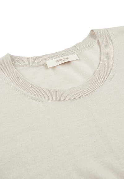 Beige gestrickter Pullover mit Rundhalsausschnitt, der eine glatte Textur und einen gerippten Kragen aufweist. Das Etikett trägt die Aufschrift "SLOWEAR" nahe dem Ausschnitt.
