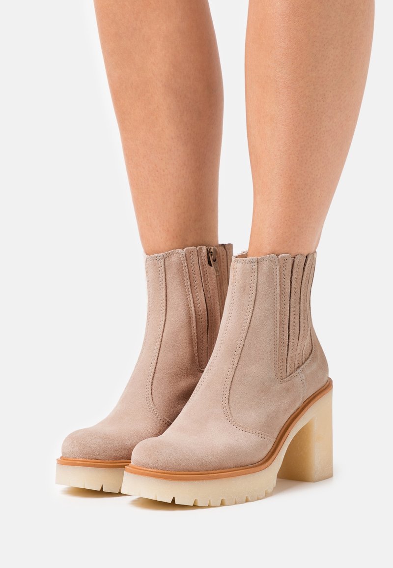 Botines de ante beige con un alto tacón de bloque, paneles laterales elásticos y una suela texturizada. Los detalles incluyen costuras cosidas y bordes ondulados.