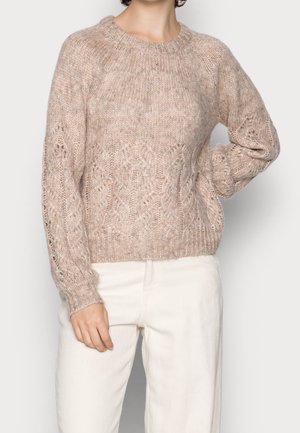 Jersey beige grueso de punto con patrones de trenzas, combinado con pantalones blancos de talle alto, mostrando un corte relajado y detalles de tela texturizada.