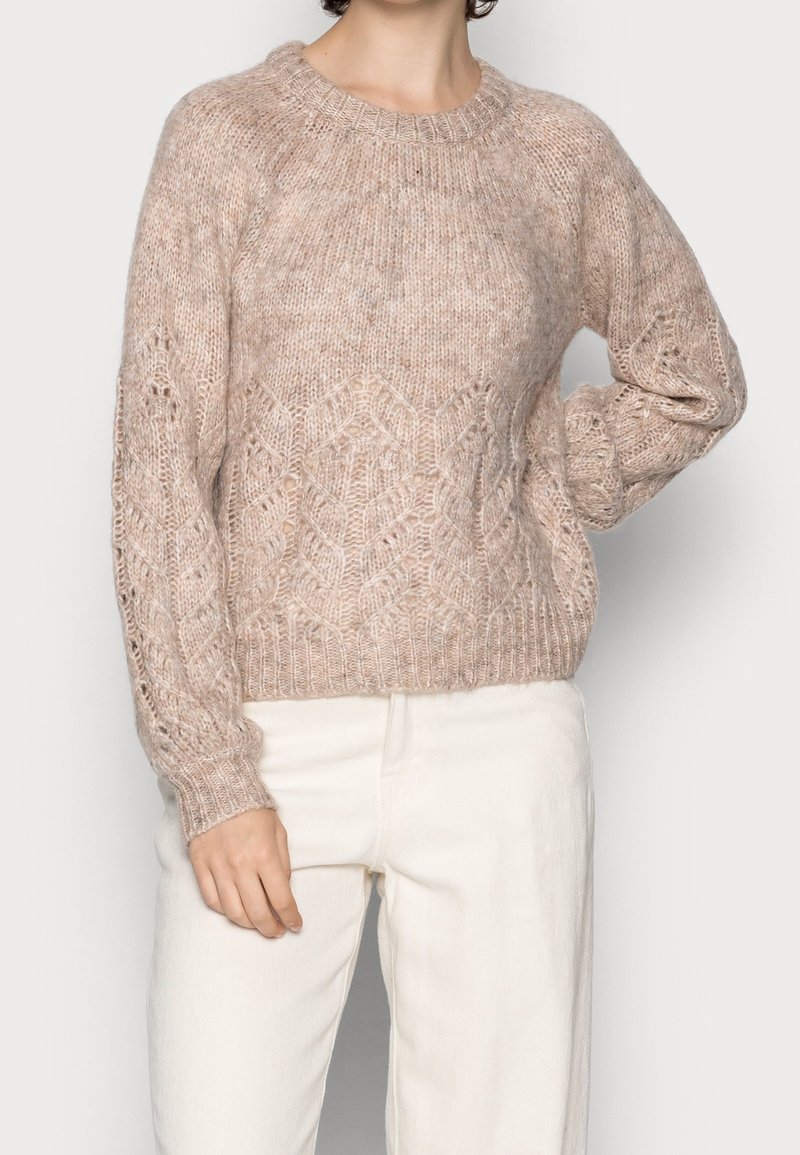 Pull beige en maille épaisse avec motifs torsadés porté avec un pantalon blanc taille haute, montrant une coupe décontractée et des détails de tissu texturé.