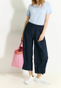 Lichtblauwe polo met korte mouwen, marineblauwe geplooide culottes en witte platform sneakers. Houdt een roze en witte geblokte gevlochten tas vast.