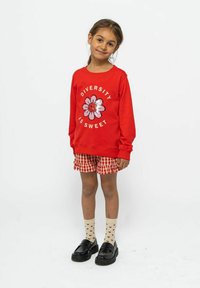 Červená mikina s nápisem "Diverzita je sladká" a květinovým vzorem, spárovaná s kostkovanými kraťasy, vzorovanými ponožkami a černými botami.