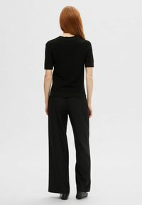 Zwarte gebreide top met korte mouwen en een ronde hals, gecombineerd met losse zwarte broeken, vanuit de achterkant gezien tegen een lichte achtergrond.